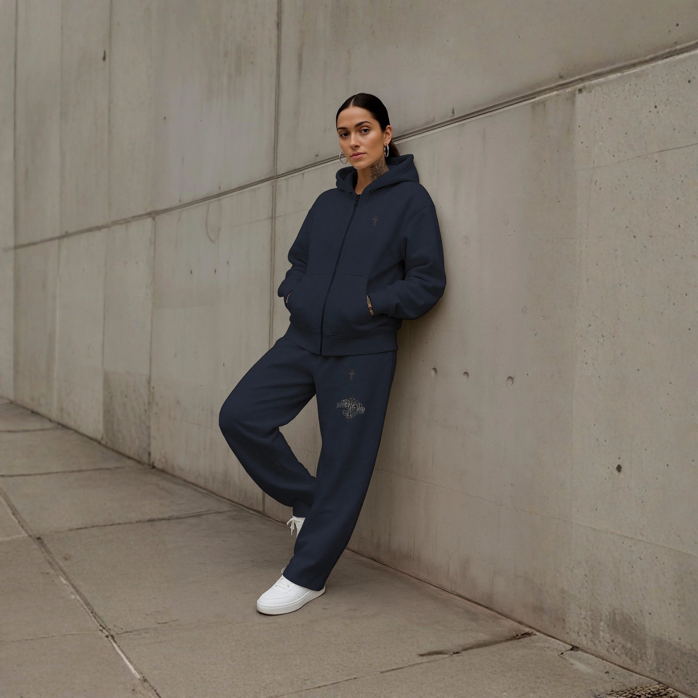 Female model wearing MAXOMclo Elysium Tracksuit NAVY – oversized heavyweight navy zip-up hoodie and baggy sweatpants with minimalist cross embroidery, premium unisex streetwear set photographed against a modern concrete wall.