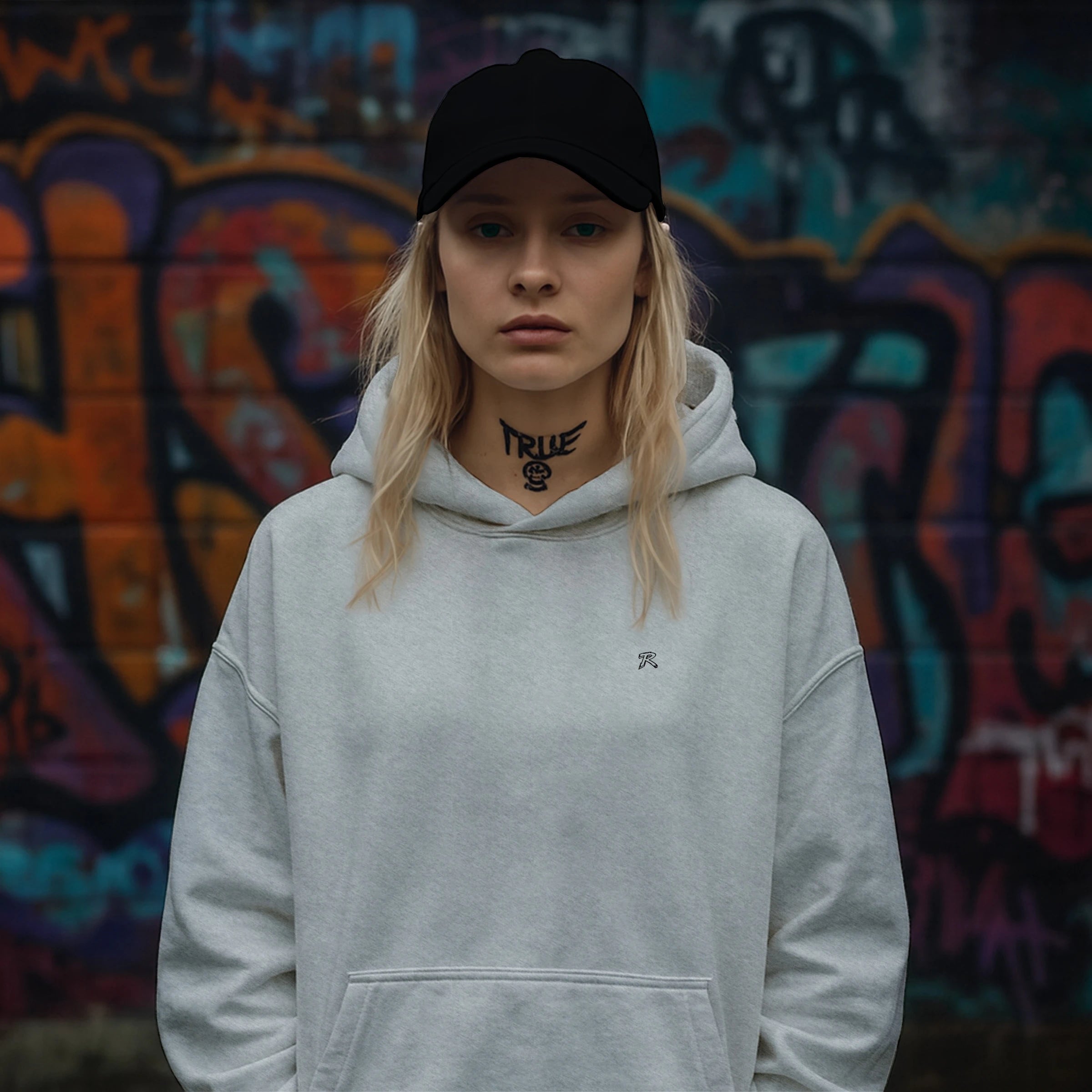 Female model wearing REMNANT. COLD CAP in black with a grey oversized minimal hoodie, posing in front of a colorful graffiti wall; low-profile unstructured cotton twill streetwear cap with subtle embroidered branding.