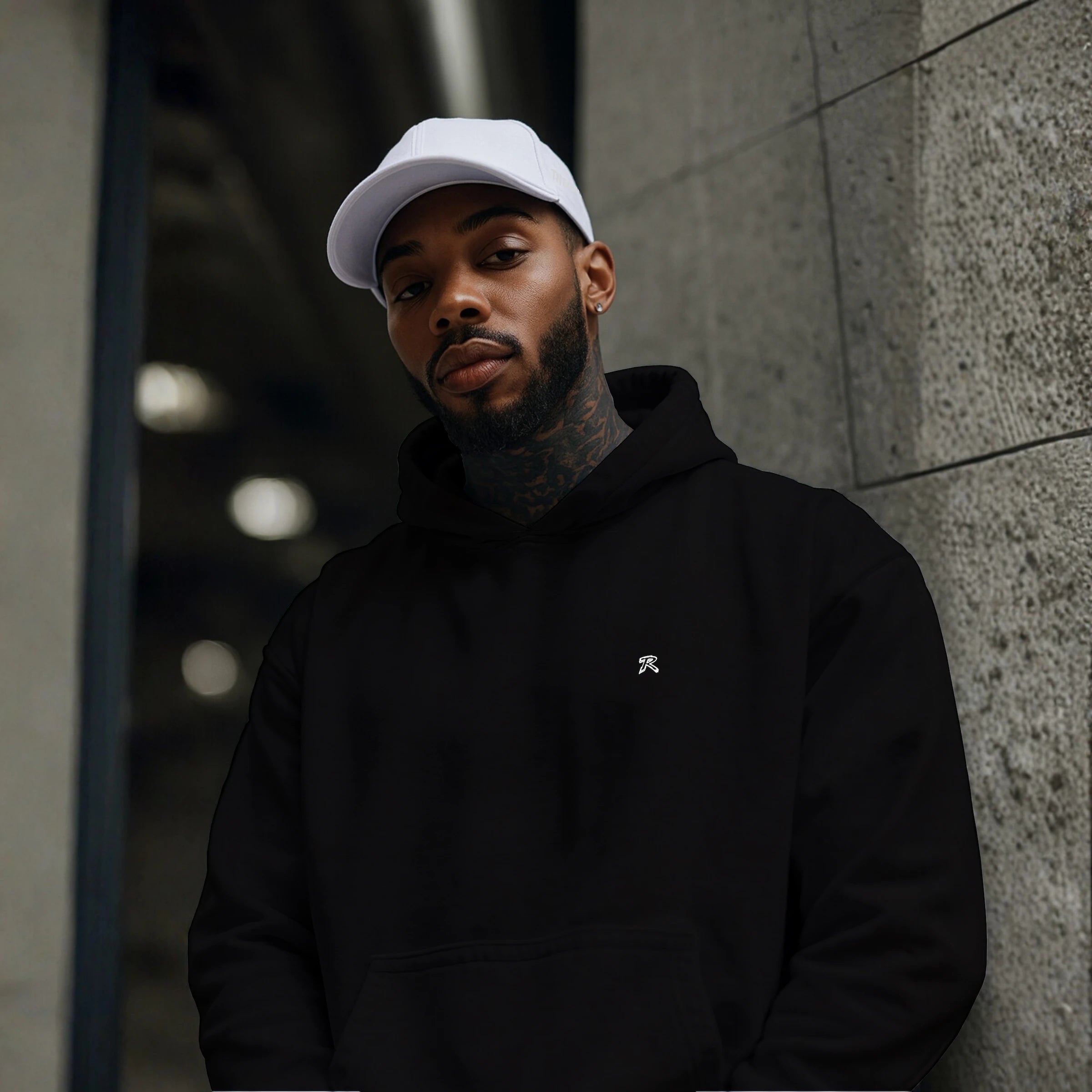 Male model wearing REMNANT. COLD CAP in white paired with a black oversized minimal hoodie, standing in an urban concrete setting; clean low-profile cotton twill streetwear cap with subtle embroidered logo for modern minimalist fashion.