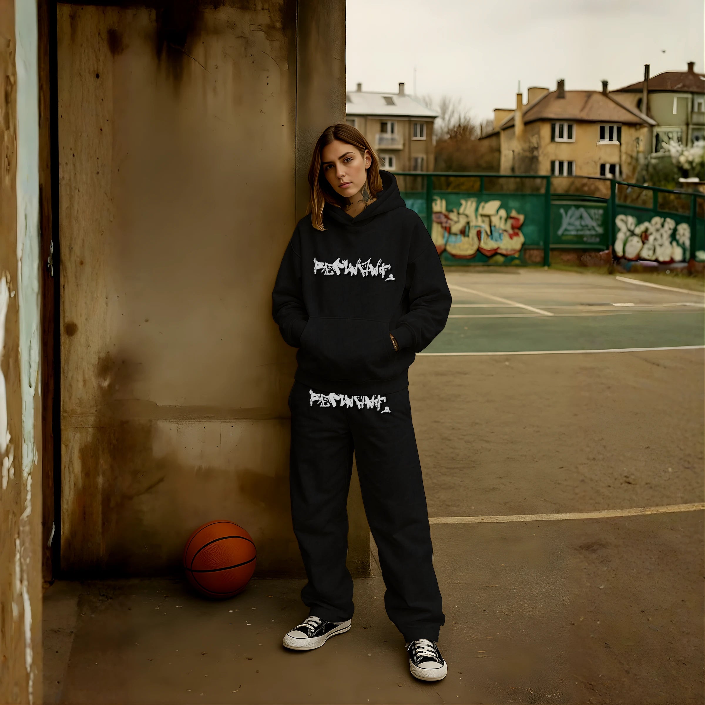 Female model wearing REMNANT DROP.ØNE oversized black graffiti streetwear set – heavyweight 380 GSM hoodie with drop-shoulder fit and matching baggy sweatpants featuring bold street-art graphics, photographed on an urban basketball court.