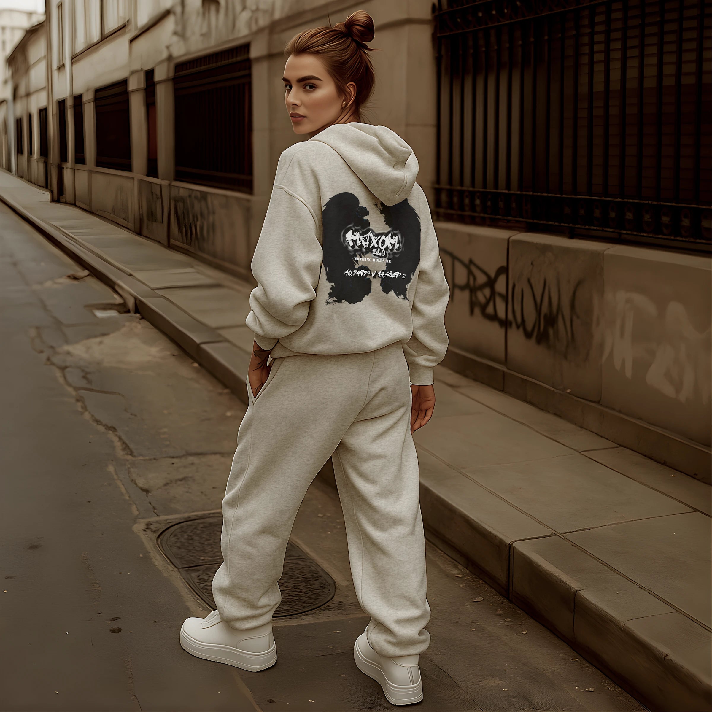 Female model wearing REMNANT. ECLIPSE_R0 oversized grey streetwear set with heavyweight hoodie featuring bold angel-wing backprint and “Nothing Holds Me” graffiti slogan, styled in an urban alley for a modern minimalist streetwear look.