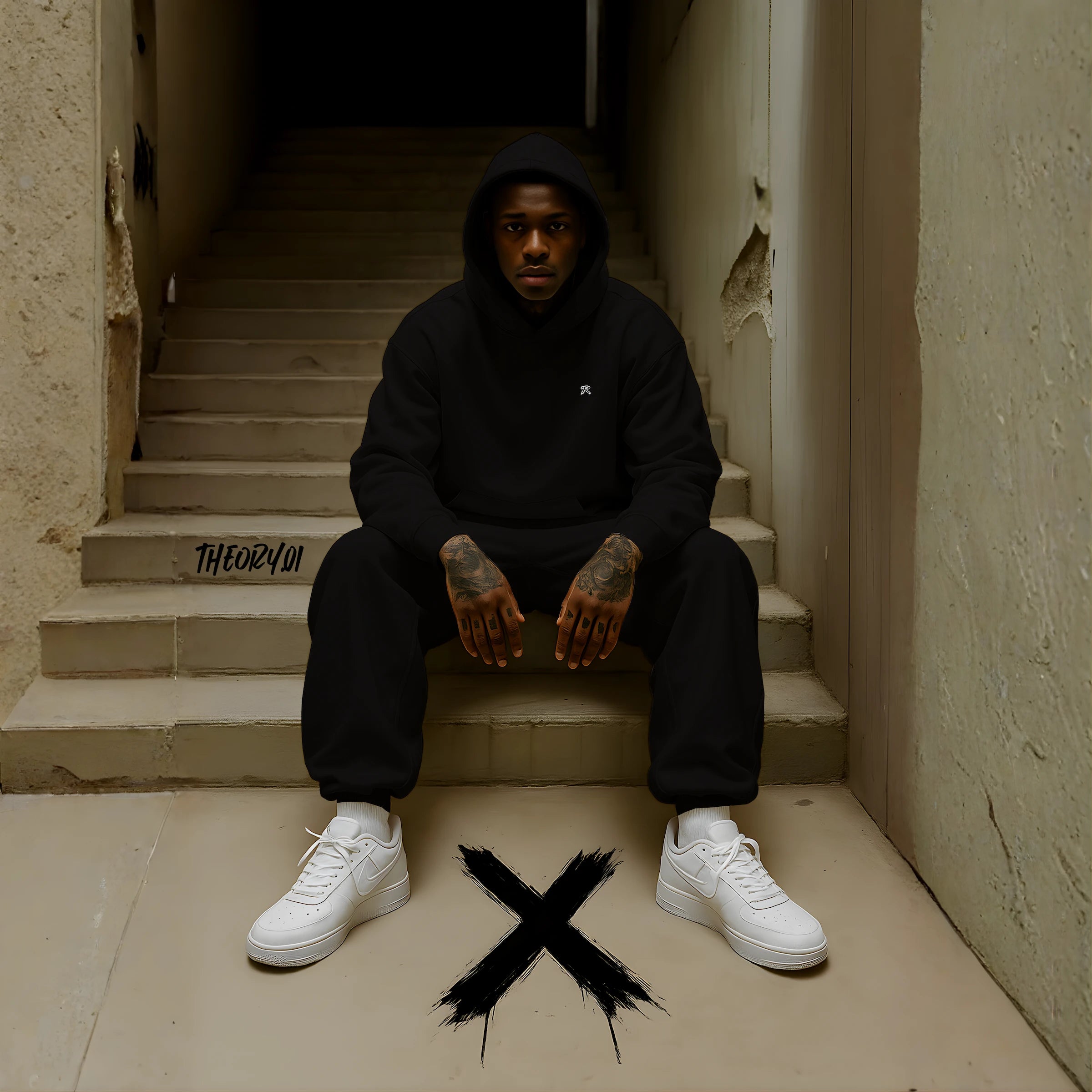 Male model wearing REMNANT. NOIR THEORY.01 oversized black fleece streetwear set with minimalist embroidered logo, sitting on concrete stairs in an urban setting with bold black X graphic on the ground, showcasing premium heavyweight hoodie and baggy sweatpants in a clean minimal aesthetic.